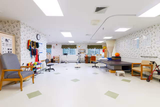 Bright, spacious rehabilitation therapy room with exercise equipment, therapy tables, chairs, and mobility aids.