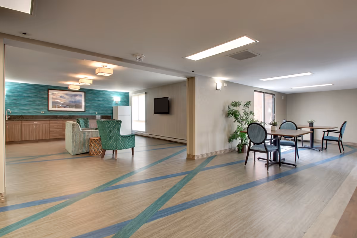 A spacious common area in a senior living facility featuring a kitchenette with wooden cabinets, a white refrigerator, and a teal accent wall with a framed picture. The room has a seating area with two green upholstered chairs and a small table, as well as a dining area with tables and chairs near large windows. The floor has a decorative pattern with blue and green stripes, and there are ceiling lights and wall sconces providing illumination.