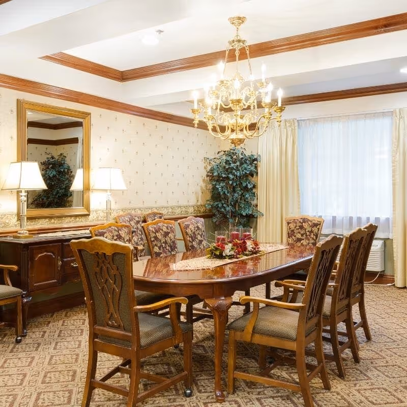 A formal dining room with a polished wooden dining table surrounded by eight upholstered wooden chairs. The room features patterned carpet, wallpaper with a decorative border, a large mirror above a wooden sideboard with two table lamps, a chandelier hanging from the ceiling, and a large window with cream-colored curtains. There is also a potted plant in the corner.
