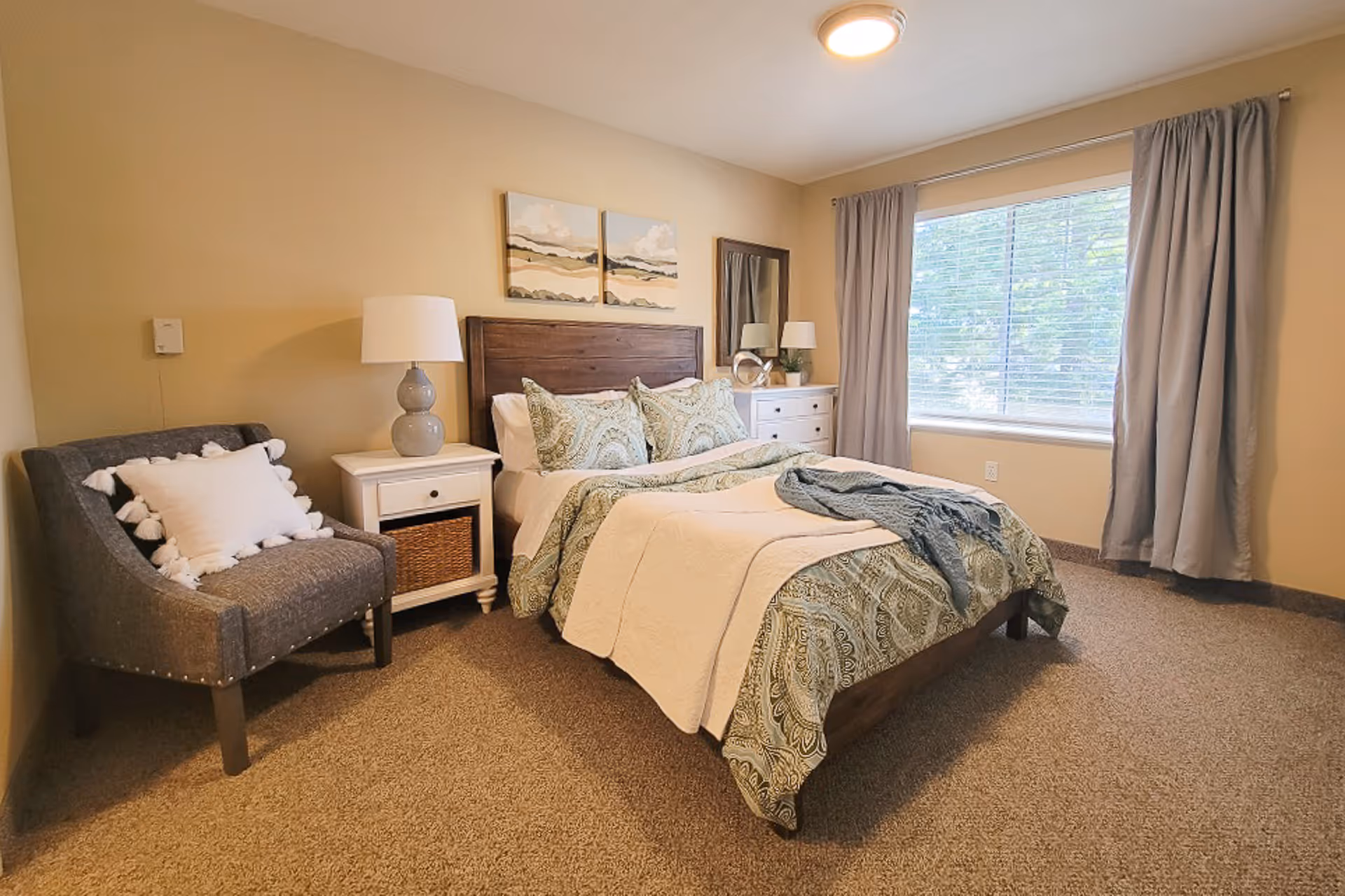 Well-lit furnished bedroom with a bed, nightstands, an upholstered chair, and a large window with curtains.