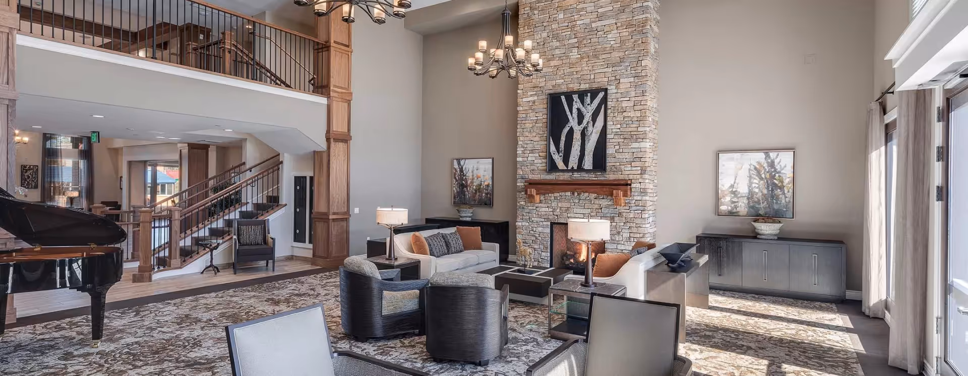Spacious senior living lounge with seating groups, a stone fireplace, grand piano, and a mezzanine staircase.