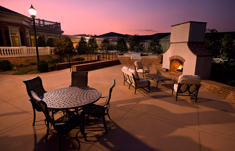 Outdoor patio area at dusk with a round metal table and four chairs in the foreground, cushioned seating arranged around a lit outdoor fireplace, brick walls, and a lamppost near a building with white railings and windows.