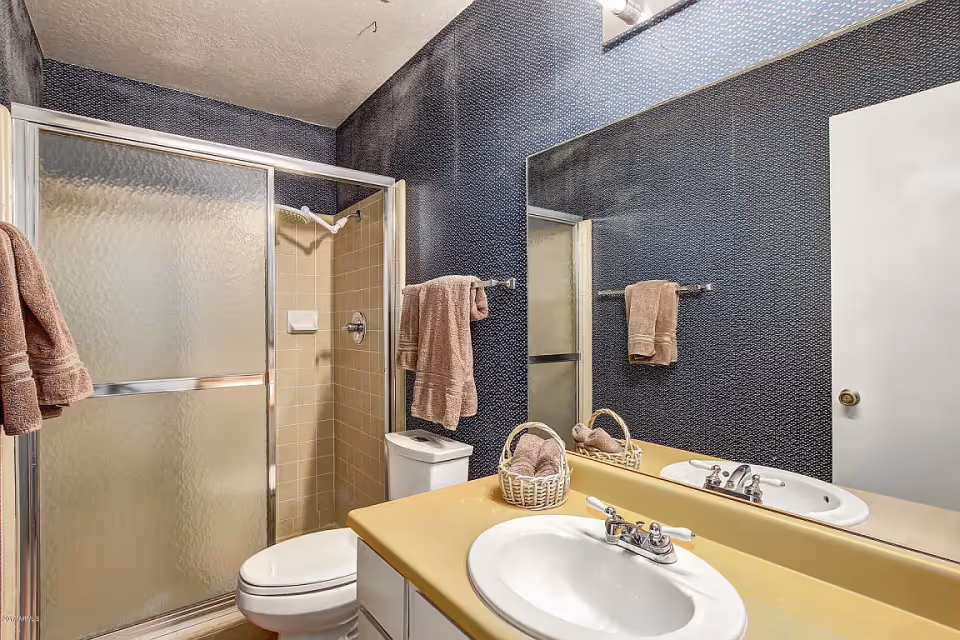 A bathroom with a beige countertop sink, a large mirror, a toilet, and a shower with frosted glass sliding doors. The walls have a dark textured wallpaper, and there are brown towels hanging on towel racks and placed in a small basket on the countertop.