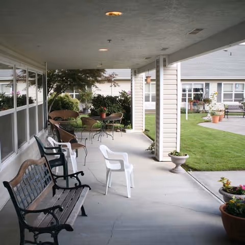 Covered outdoor patio area with various seating options including wooden benches, plastic chairs, and metal tables with chairs. The patio overlooks a well-maintained grassy area with potted plants and shrubs near a building with multiple windows.