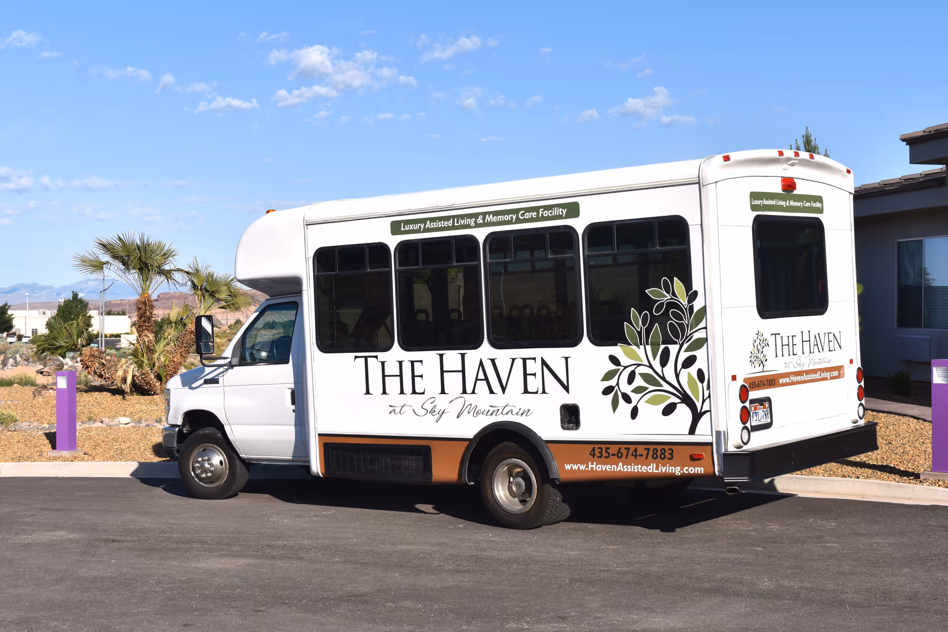 A white shuttle bus parked outside on a sunny day with clear blue sky. The bus has signage for The Haven at Sky Mountain Independent and Assisted Living Community, including a tree logo and contact information. There are palm trees and desert landscaping in the background.
