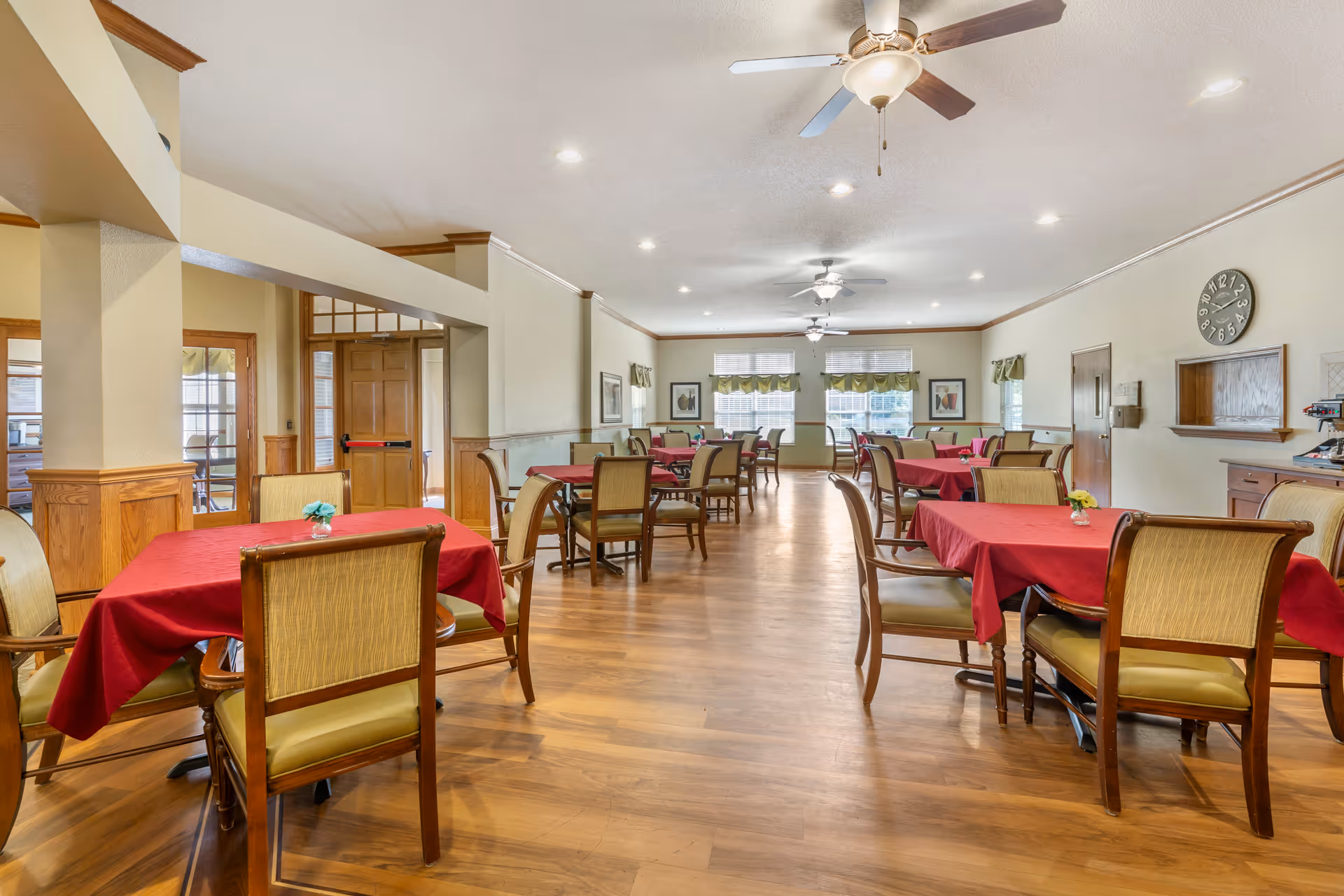 A spacious dining room with wooden floors and multiple tables covered with red tablecloths. Each table is surrounded by wooden chairs with cushioned seats. The room has ceiling fans with lights, large windows with green valances, and framed artwork on the walls. There is a clock on the wall and a small service window area with a coffee machine on the right side.