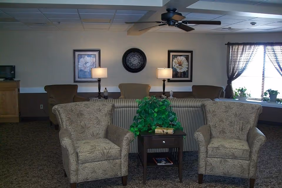 Communal living room with two patterned armchairs flanking a small table topped with a potted plant, a striped sofa, lamps, wall clock and framed artwork near a window with curtains.