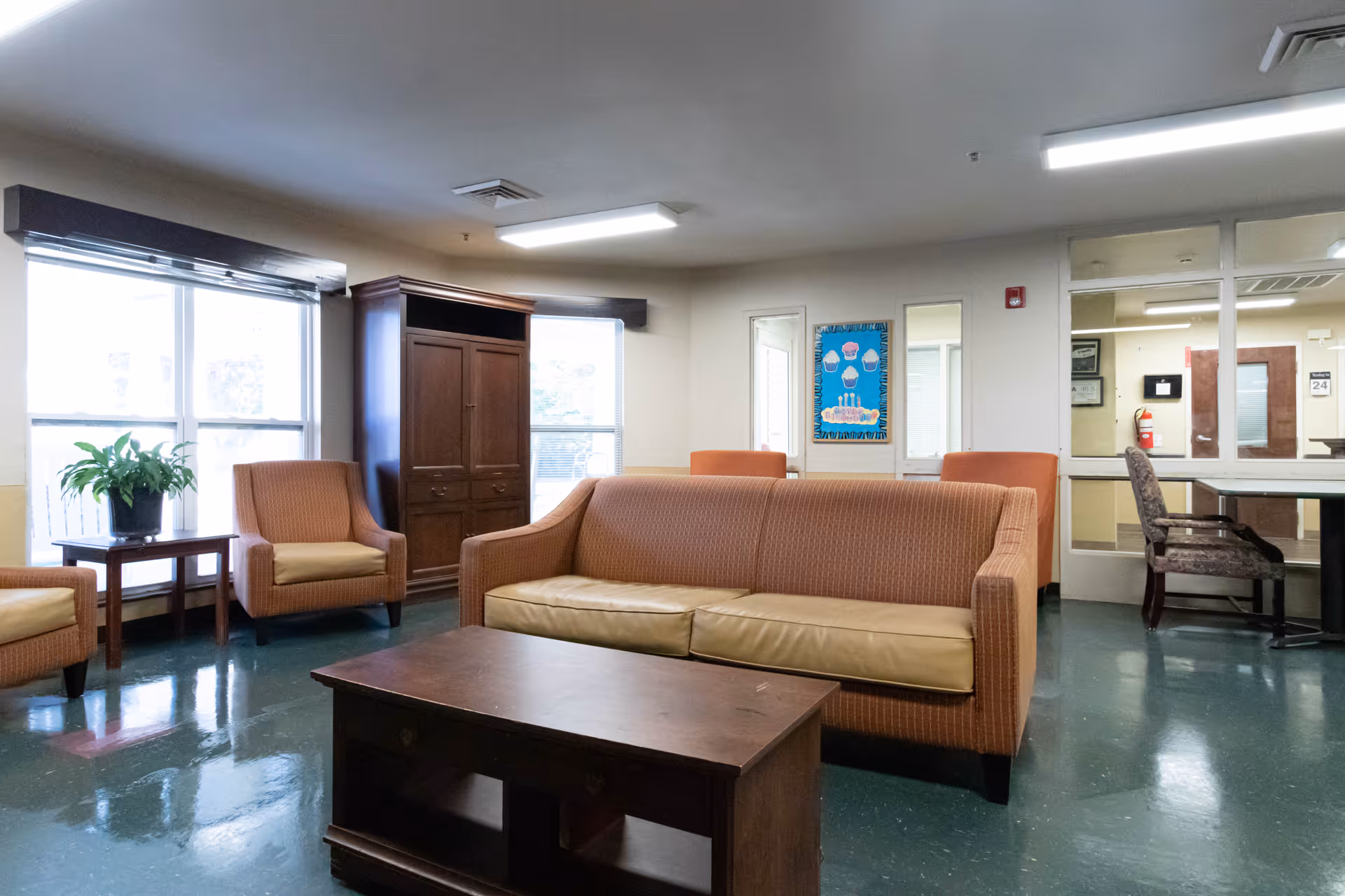 A common area with orange cushioned seating including a sofa and armchairs, a wooden coffee table, a wooden cabinet, and large windows letting in natural light. There is a plant on a side table near the window and a bulletin board on the wall. The floor is green and shiny, and fluorescent lights illuminate the room.
