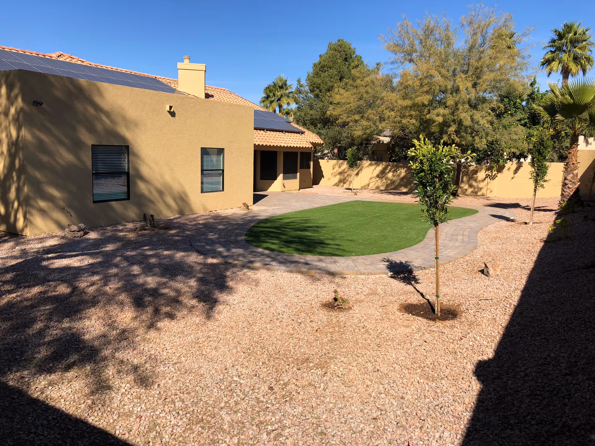 A backyard area with a beige stucco building featuring two windows and a tiled roof with solar panels. The yard has a small patch of green artificial grass surrounded by a stone pathway, with gravel covering the rest of the ground. Several small trees and bushes are planted around the yard, and there are taller trees and palm trees in the background under a clear blue sky.