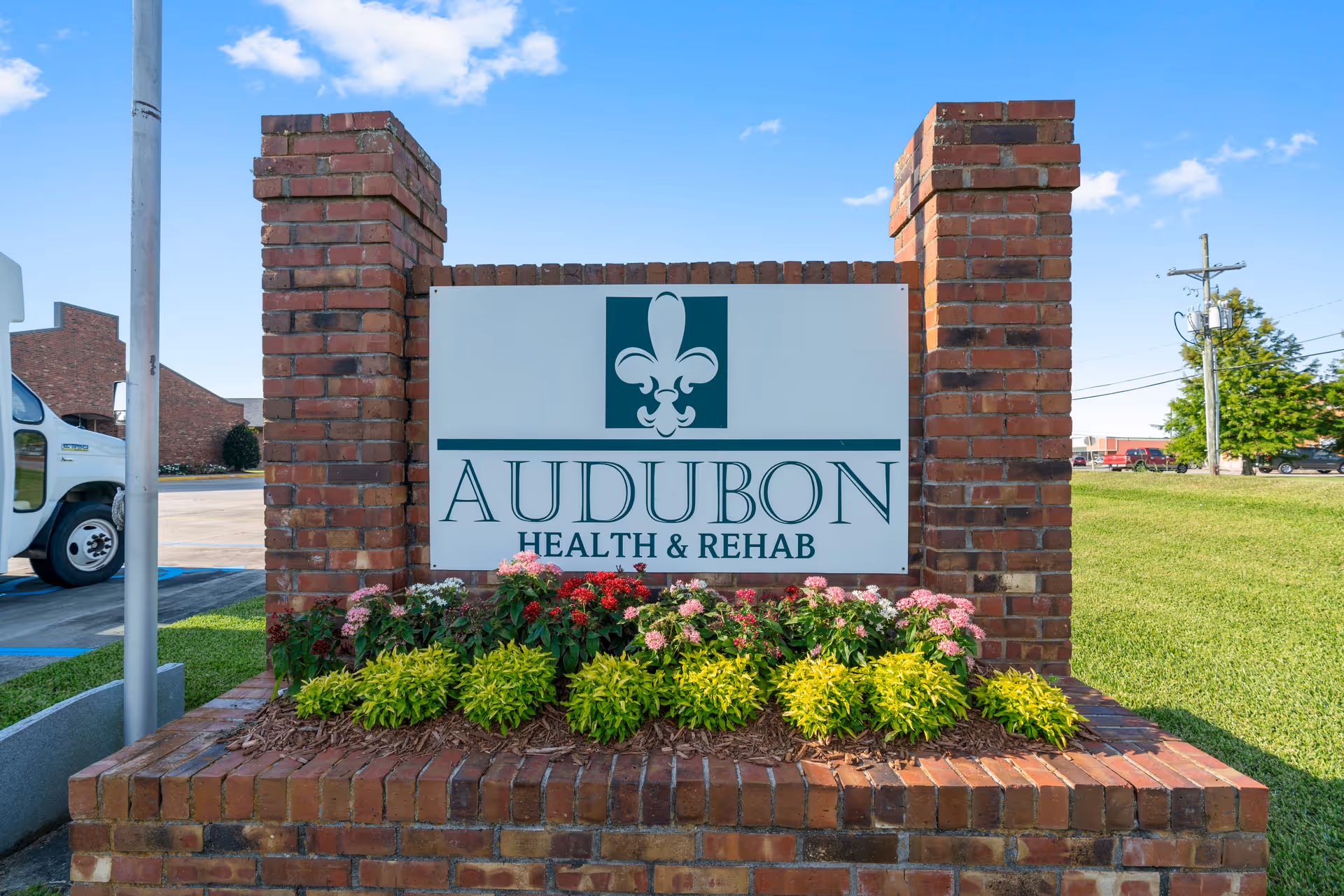A brick sign for Audubon Health & Rehab with a green and white logo featuring a fleur-de-lis. The sign is surrounded by a flower bed with various colorful flowers and green plants. The background shows a parking lot, a white vehicle, grassy area, and a clear blue sky with some clouds.