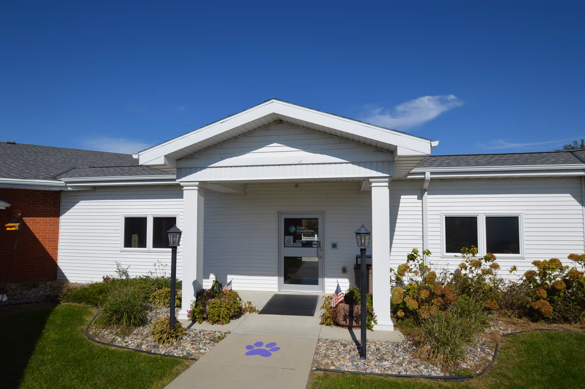 Front entrance of a single-story white building with a covered porch supported by two columns. There are two black lamp posts on either side of the walkway leading to the door, which has a glass panel. The building is surrounded by small bushes and a well-maintained lawn under a clear blue sky.