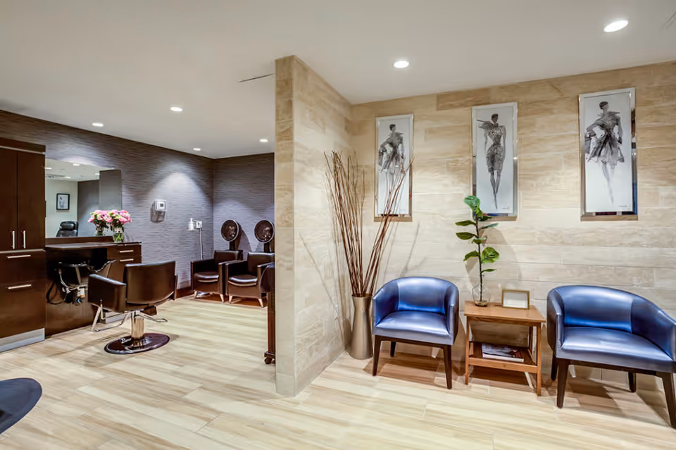 Interior view of a stylish salon area with wooden flooring and beige tiled walls. The space features two blue leather chairs with a small wooden table between them, decorated with a plant and framed pictures on the wall above. To the left, there are salon chairs, hair drying stations, and a counter with a vase of pink flowers.
