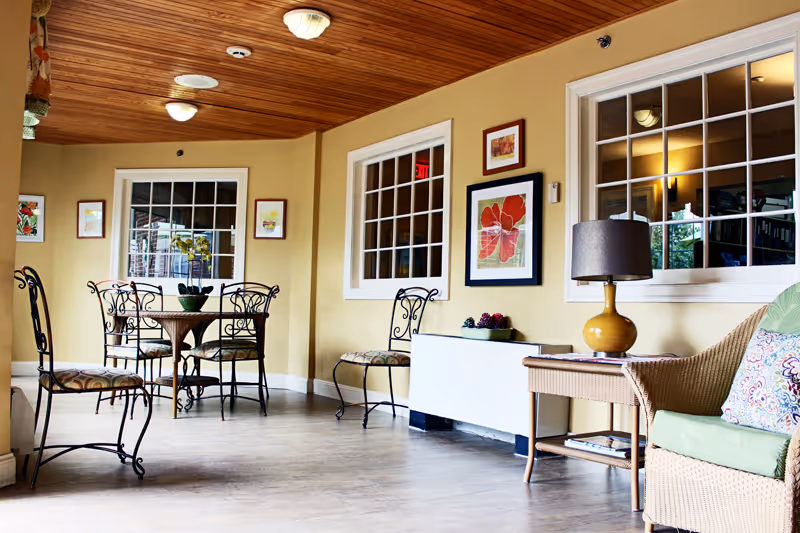 Sunlit common room with a wicker sofa, round dining table and chairs, artwork on the walls and large windows.