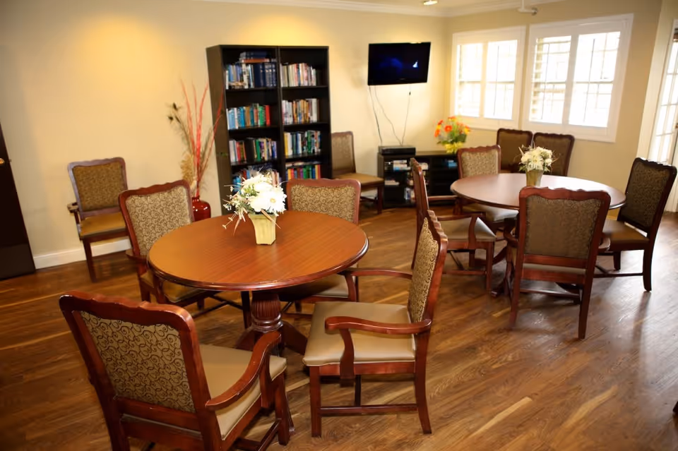 A cozy common area with two round wooden tables, each surrounded by six upholstered chairs. Each table has a small floral centerpiece. In the background, there is a black bookshelf filled with books, a flat-screen TV mounted on the wall, and a window with white shutters letting in natural light. The room has wooden flooring and a warm, inviting atmosphere.