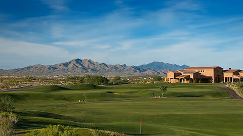 A wide view of a green golf course with a red flag on the putting green, surrounded by desert vegetation. In the background, there is a large building with a southwestern architectural style and mountains under a blue sky with some clouds.