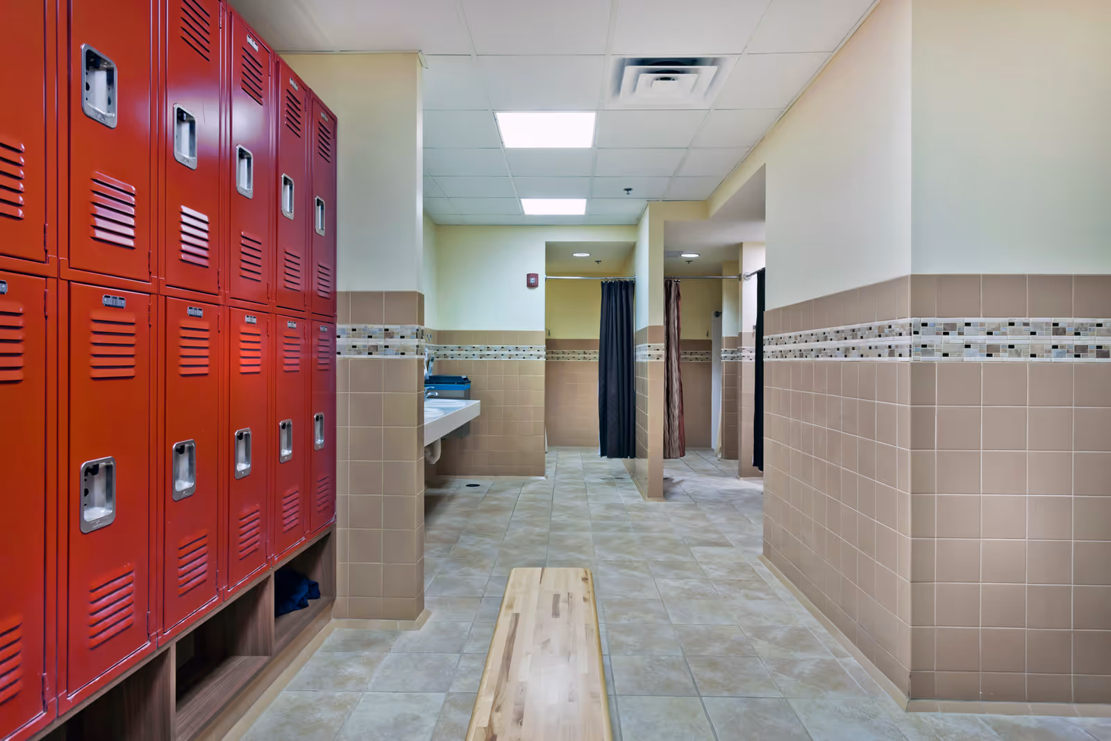 Tiled locker room featuring red lockers, a wooden bench, sinks, and shower stalls with curtains.