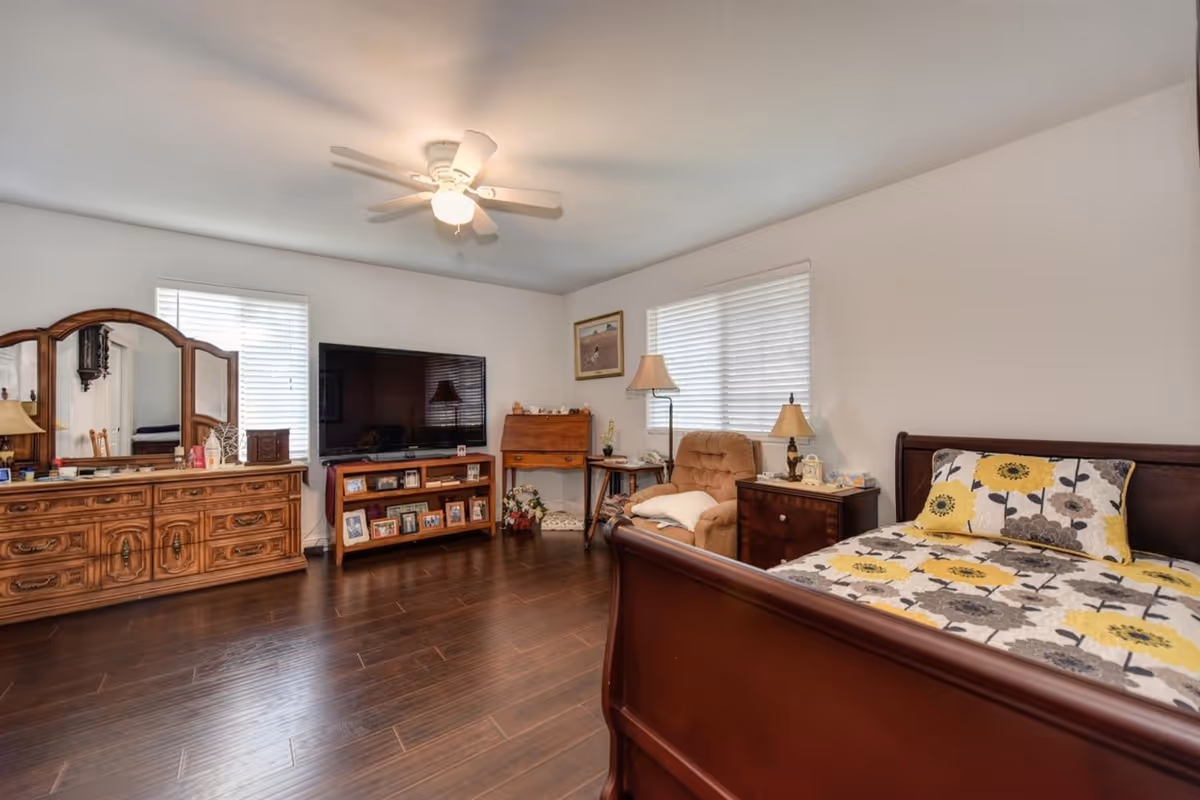 Well-lit bedroom with dark hardwood floors, a wooden bed with floral bedding, dresser with mirror, TV on a stand, and a recliner with bedside tables.