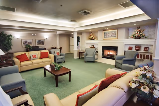 A cozy living room in an assisted living facility featuring beige sofas with red and patterned cushions, several blue armchairs, a wooden coffee table with a plant centerpiece, a fireplace with a fire burning, decorative flowers, framed pictures on the walls, and a green carpeted floor.