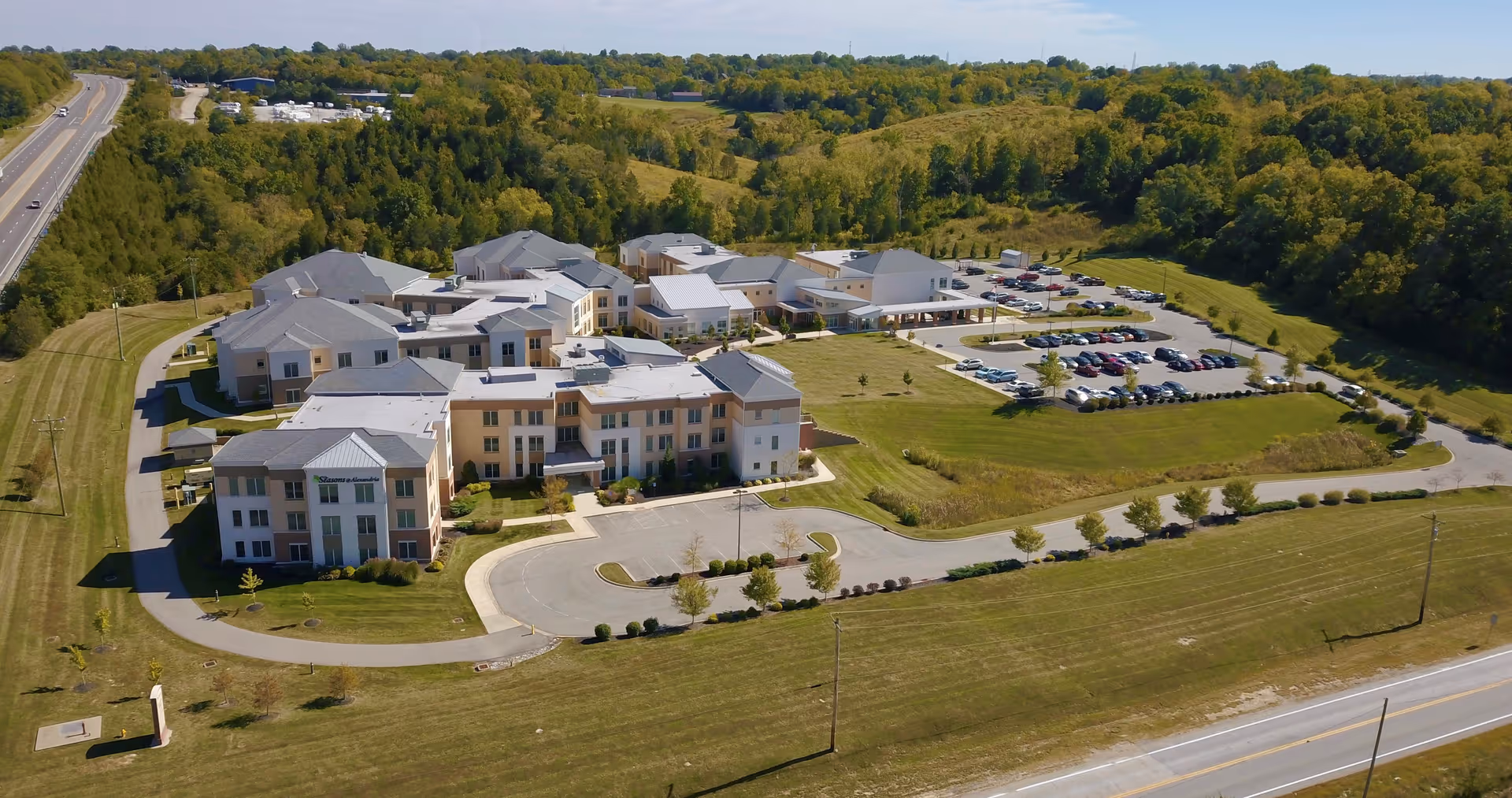 Aerial view of The Seasons@Alexandria - a BLC community, showing multiple connected buildings surrounded by green lawns and trees, with parking lots and a road nearby under a clear sky.