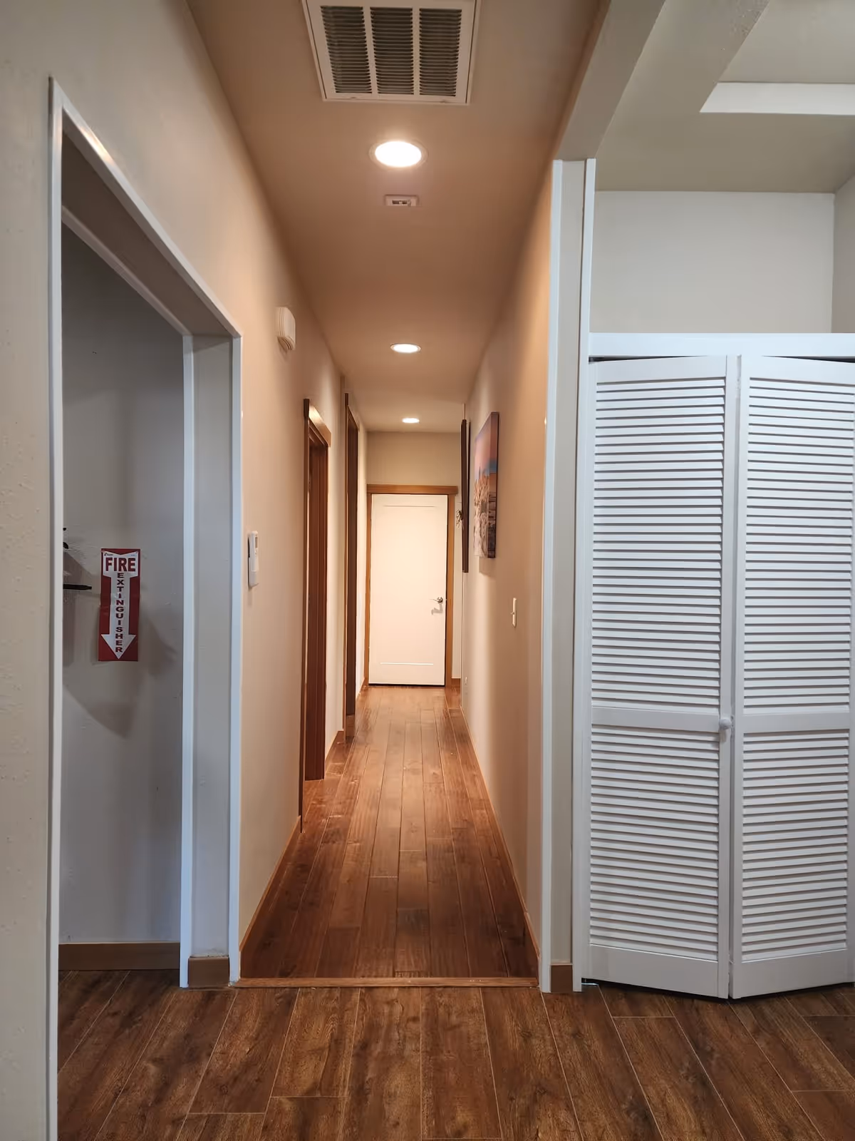 A long indoor hallway with wooden flooring and beige walls, illuminated by ceiling lights. On the left side, there is an open doorway with a fire extinguisher sign visible inside. On the right side, there is a white folding door. At the end of the hallway, there is a closed white door and a framed picture on the right wall.
