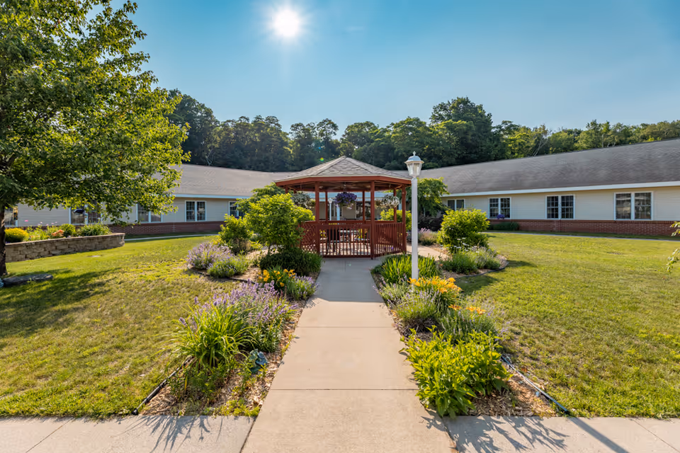 Landscaped courtyard with a central wooden gazebo, paved path, flower beds and a surrounding single-story building under a sunny sky.
