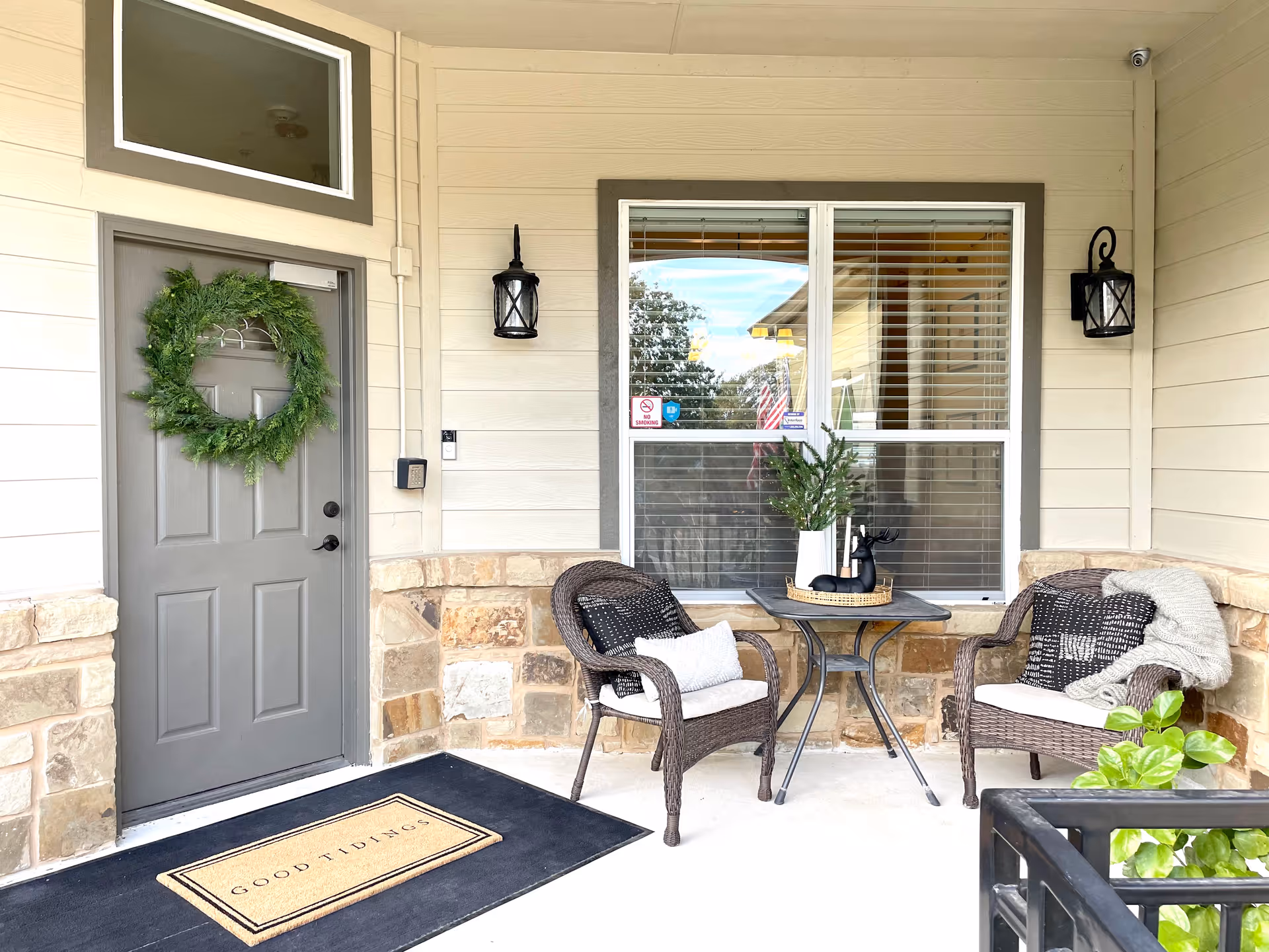 Covered front porch with a gray door adorned with a wreath, two wicker chairs and a small table on a welcome mat.