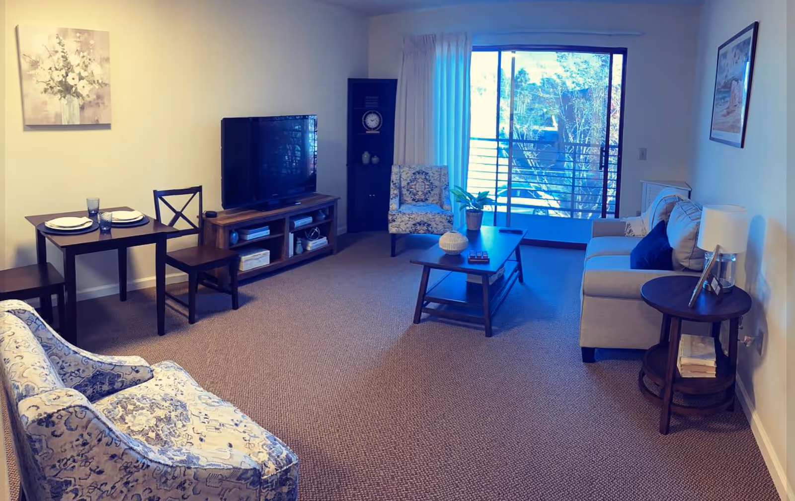 A cozy living room with a beige sofa, two patterned armchairs, a wooden coffee table with decorative items, a TV on a wooden stand, a small dining table set for two, and a sliding glass door leading to a balcony with a view of trees outside.