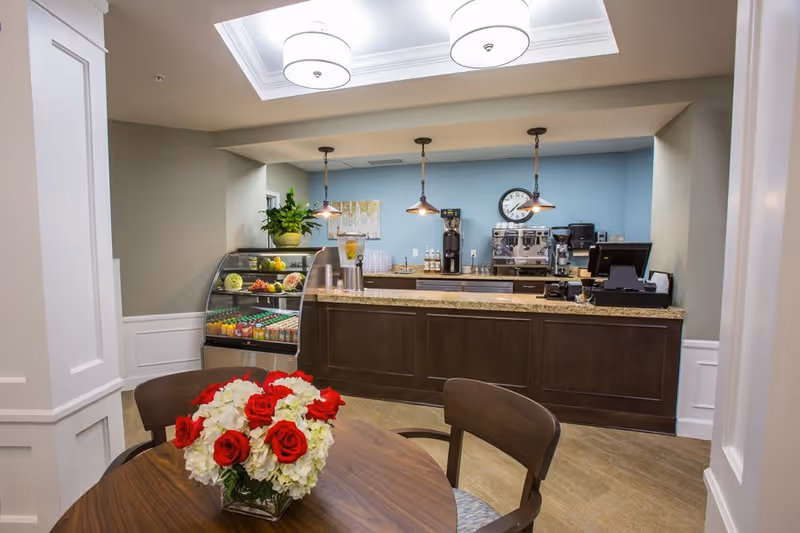 A small cafe or refreshment area with a wooden table and chairs in the foreground, decorated with a vase of red and white flowers. Behind the table is a counter with a granite top, coffee machines, a clock on a blue wall, and a refrigerated display case with drinks and fruit. The area is well-lit with ceiling lights and pendant lamps.