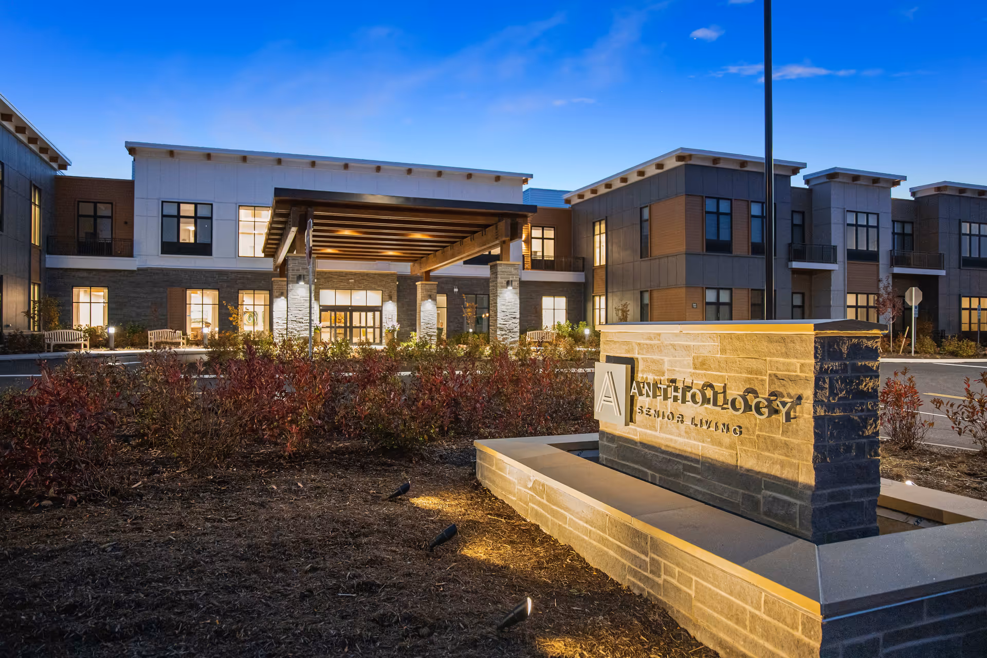 Exterior view of a modern senior living facility building at dusk with illuminated windows and a covered entrance. In the foreground, there is a stone sign with the text 'Anthology Senior Living' surrounded by landscaping and small lights.