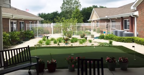 Outdoor courtyard area at RidgeCrest Health Campus featuring a small putting green, potted plants, benches, and surrounded by brick buildings with white fencing and greenery in the background.