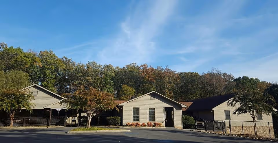 Exterior view of a single-story building with beige siding and a brown roof, surrounded by trees with green and autumn-colored leaves under a blue sky with some clouds. There is a paved parking area in front and a fenced area on the right side.