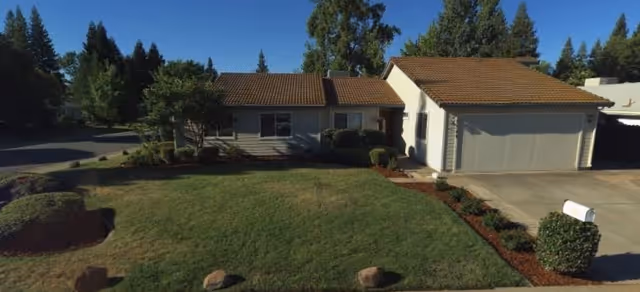 Single-story house with a tiled roof, a two-car garage, and a well-maintained front lawn with shrubs and trees under a clear sky.