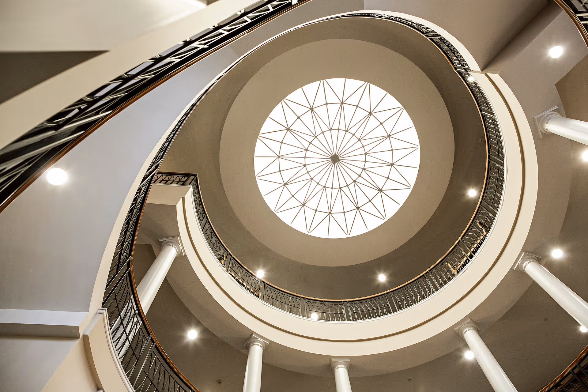 View looking up at a circular atrium with multiple levels of balconies and white columns, topped by a decorative skylight with a geometric pattern.