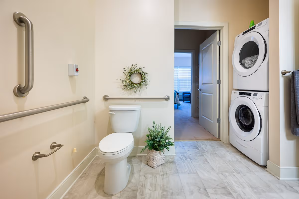 A bathroom with a white toilet, stainless steel grab bars on the walls, a small decorative wreath above the toilet, and a potted plant on the floor. To the right, there is a stacked washer and dryer unit. The bathroom opens into a carpeted room with a window and furniture visible in the background.
