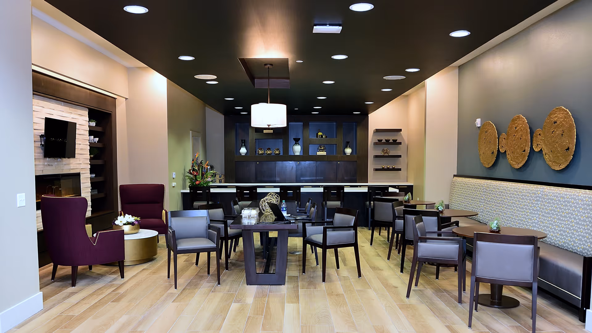 A modern senior living common area with wooden flooring, multiple tables and chairs arranged for seating, a long cushioned bench along one wall, decorative wall art, a fireplace with a mounted TV, and shelves with vases and decor items. The ceiling is dark with recessed lighting and a central hanging light fixture.