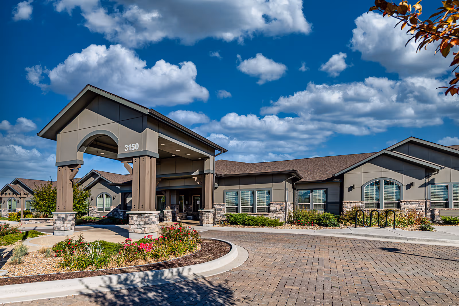 Exterior front view of a single-story senior living facility building with a covered entrance, stone and siding facade, landscaped flower beds, and a clear blue sky with scattered clouds.