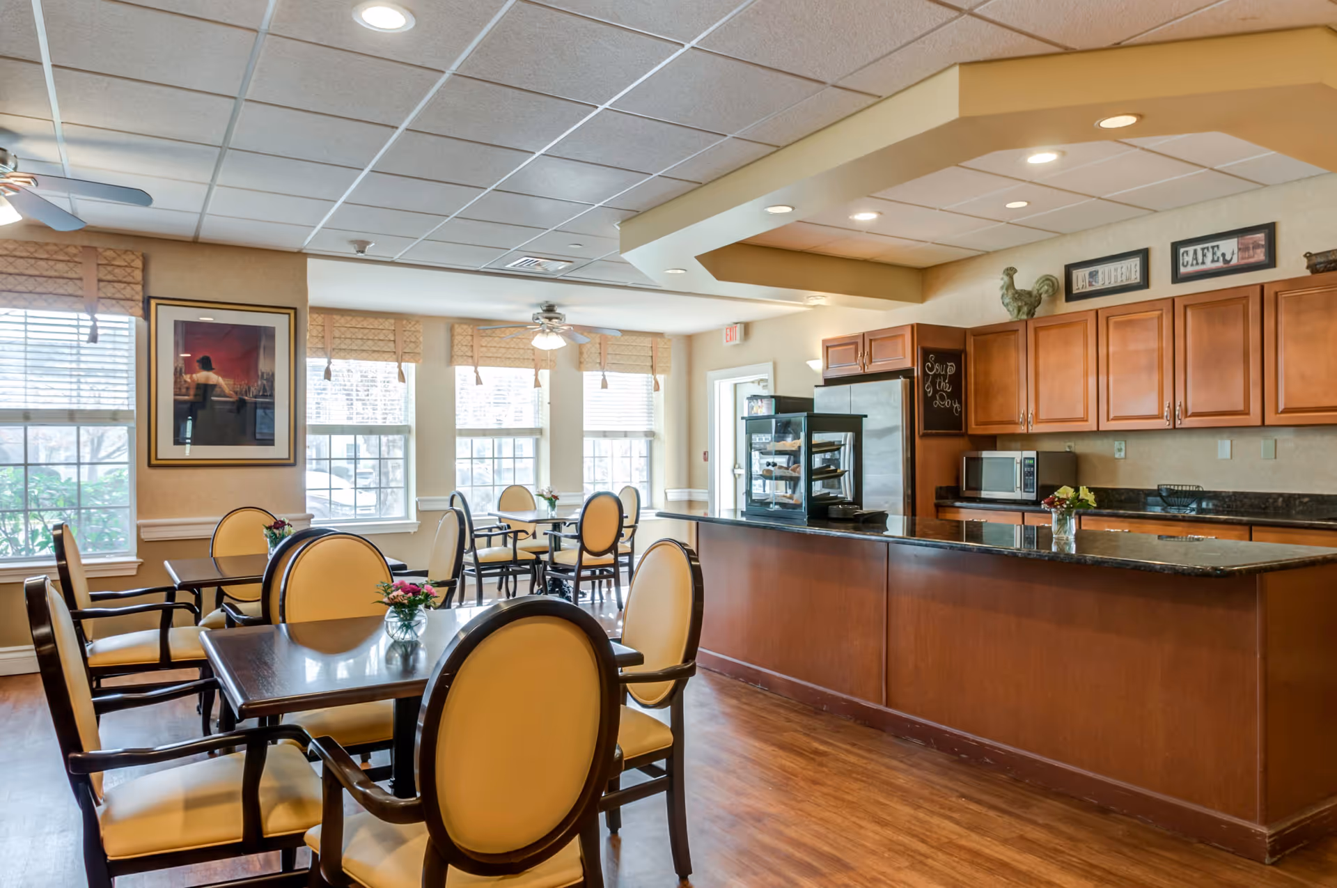 Well-lit dining area with multiple tables and upholstered chairs and a service counter with wooden cabinets and appliances.
