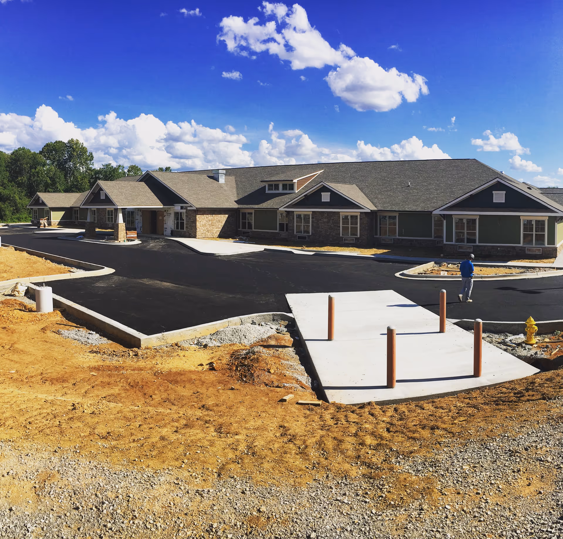 Exterior view of a newly constructed single-story senior living facility building with a paved driveway and a person walking near the entrance under a blue sky with scattered clouds.