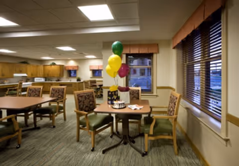 Bright dining area with tables and chairs, a small table decorated with colorful balloons, and a kitchen area in the background.