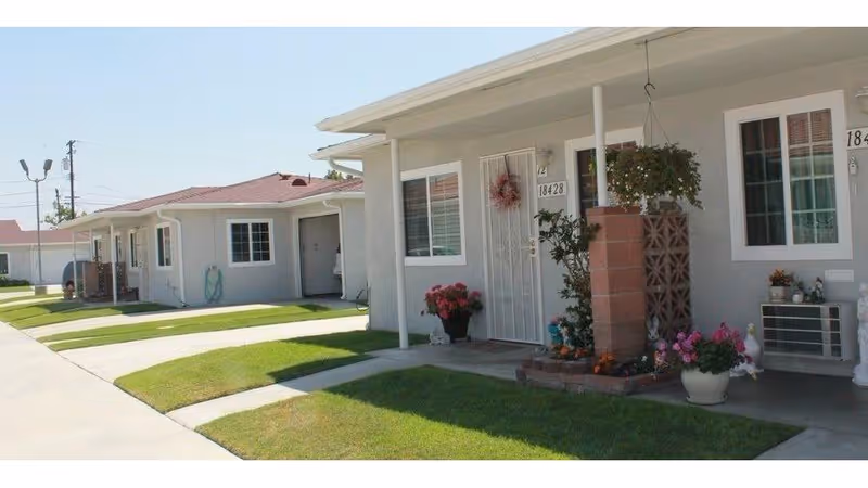 Front entrances of single-story residential units with small porches, potted plants, and manicured lawns along a sidewalk.