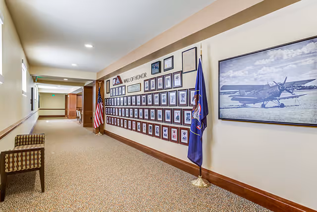 A well-lit hallway in a senior living facility with a carpeted floor and beige walls. On the right wall, there is a 'Wall of Honor' display featuring numerous framed photographs and certificates, flanked by an American flag and another flag. A large framed black and white photo of an airplane is also visible on the wall. Two patterned chairs are placed along the left wall.