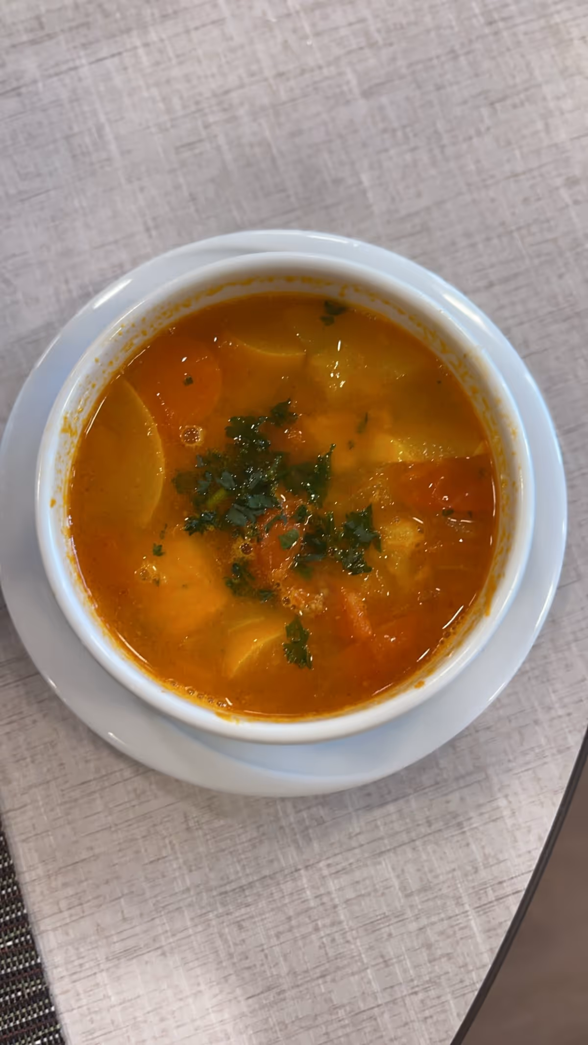 A bowl of vegetable soup garnished with chopped herbs, placed on a white saucer on a light-colored textured tablecloth.