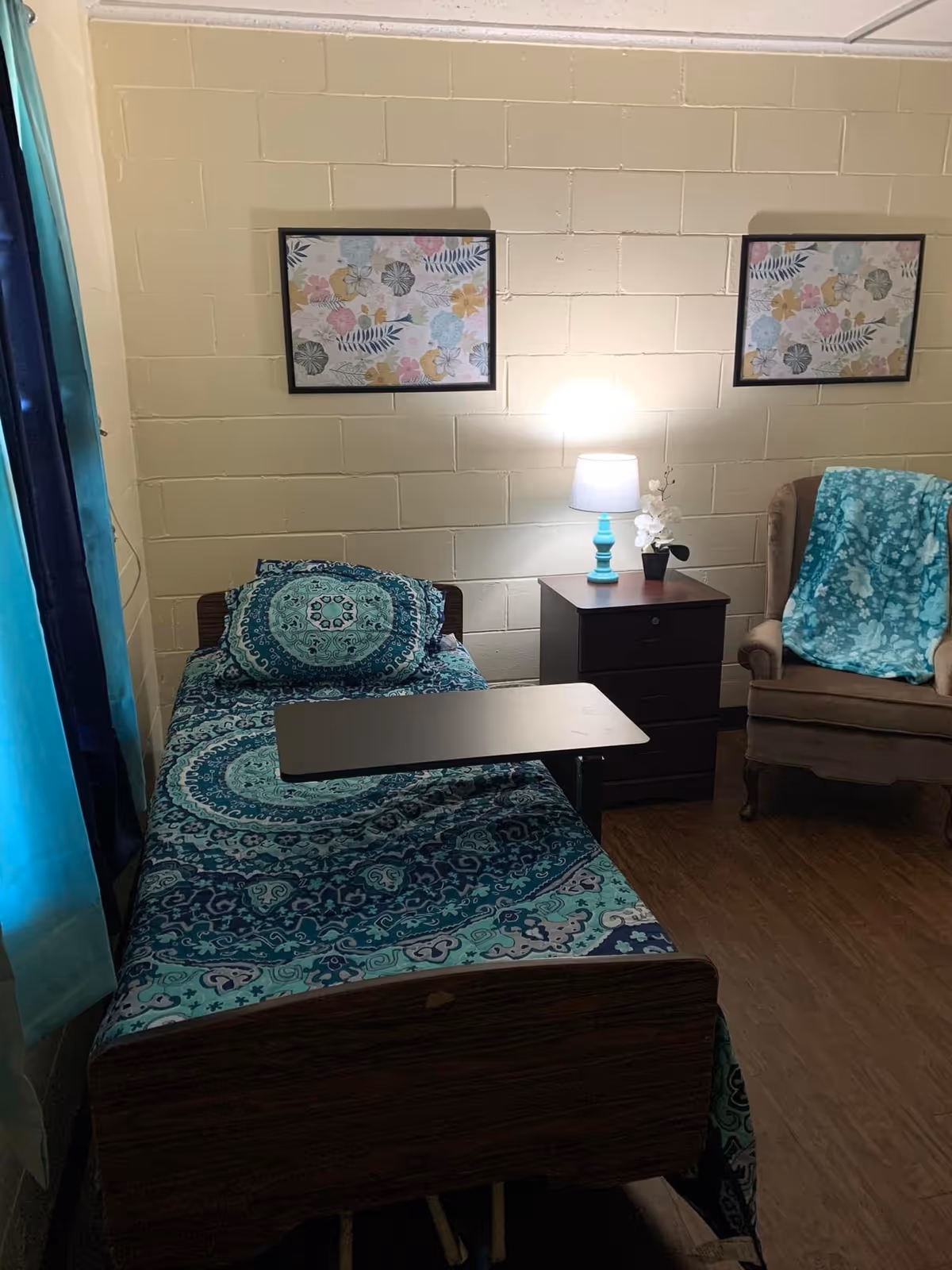 A small bedroom with a single bed covered in blue and teal patterned bedding. There is a dark wooden nightstand with a turquoise lamp and a small white flower arrangement. Next to the nightstand is a brown upholstered armchair with a blue floral blanket draped over it. Two framed floral prints hang on the beige brick wall above the bed and nightstand. A window with blue curtains is partially visible on the left side.