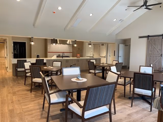 A spacious dining room with multiple square tables and cushioned chairs arranged neatly. The room features a high ceiling with exposed beams and ceiling fans. In the background, there is a kitchen area with a long counter, pendant lights, and modern cabinetry. The floor is wooden, and the walls are painted in neutral tones, creating a warm and inviting atmosphere.