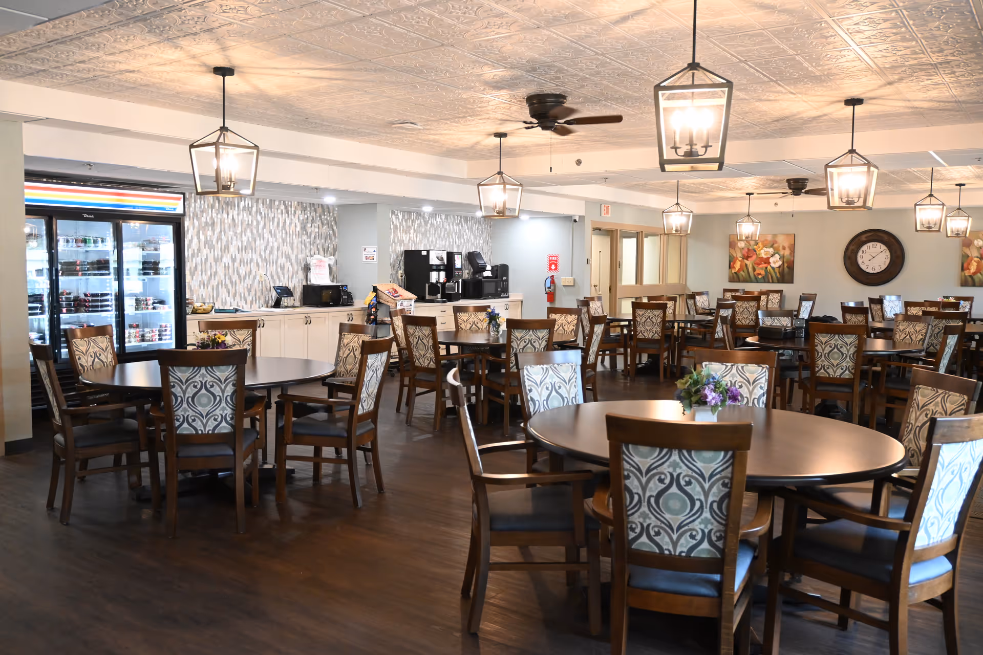 A spacious dining room with multiple round tables and wooden chairs featuring patterned upholstery. The room has a dark wood floor, pendant lights hanging from a decorative ceiling, and ceiling fans. In the background, there is a counter area with coffee machines, a microwave, and a refrigerator stocked with beverages. The walls are decorated with floral paintings and a large clock.