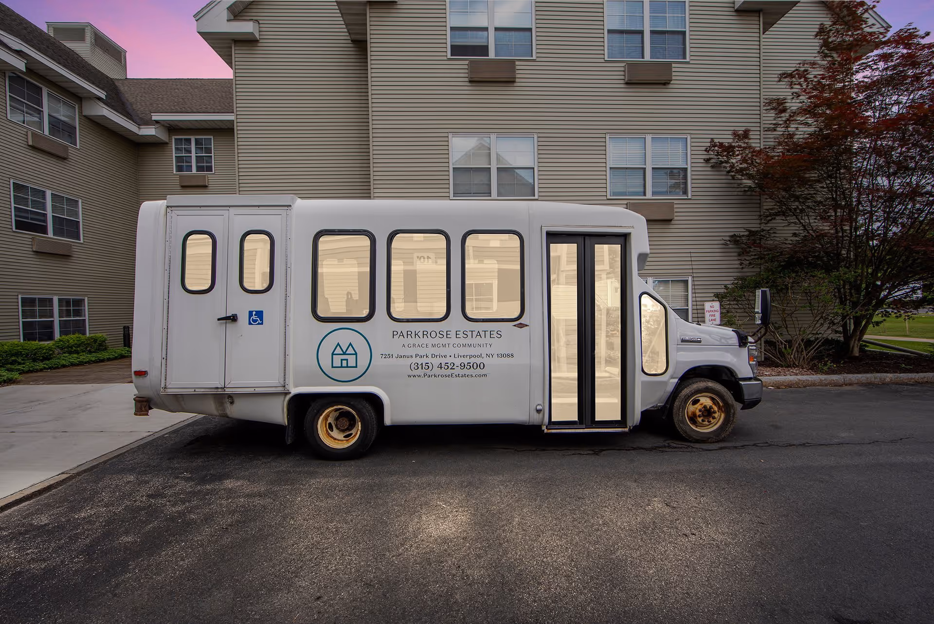 A white shuttle bus parked in front of a beige multi-story building during dusk. The bus has the logo and contact information for Parkrose Estates, a senior living community, displayed on its side. The building has multiple windows and some greenery around it.