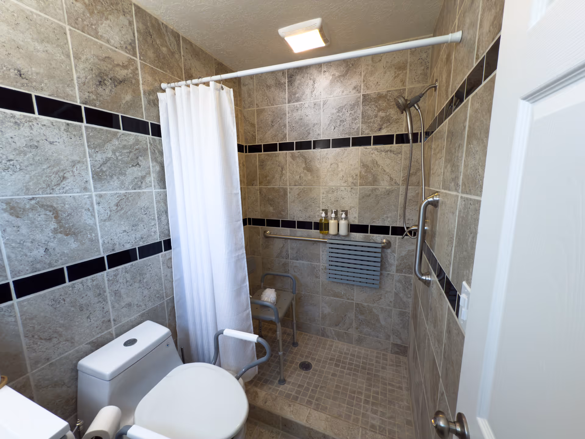 A bathroom with beige and gray tiled walls and floor. There is a white toilet with a safety rail, a walk-in shower with a white curtain, a shower chair, a grab bar, and three bottles of soap or shampoo on a shelf. The shower has a handheld showerhead and a towel hanging on the grab bar.