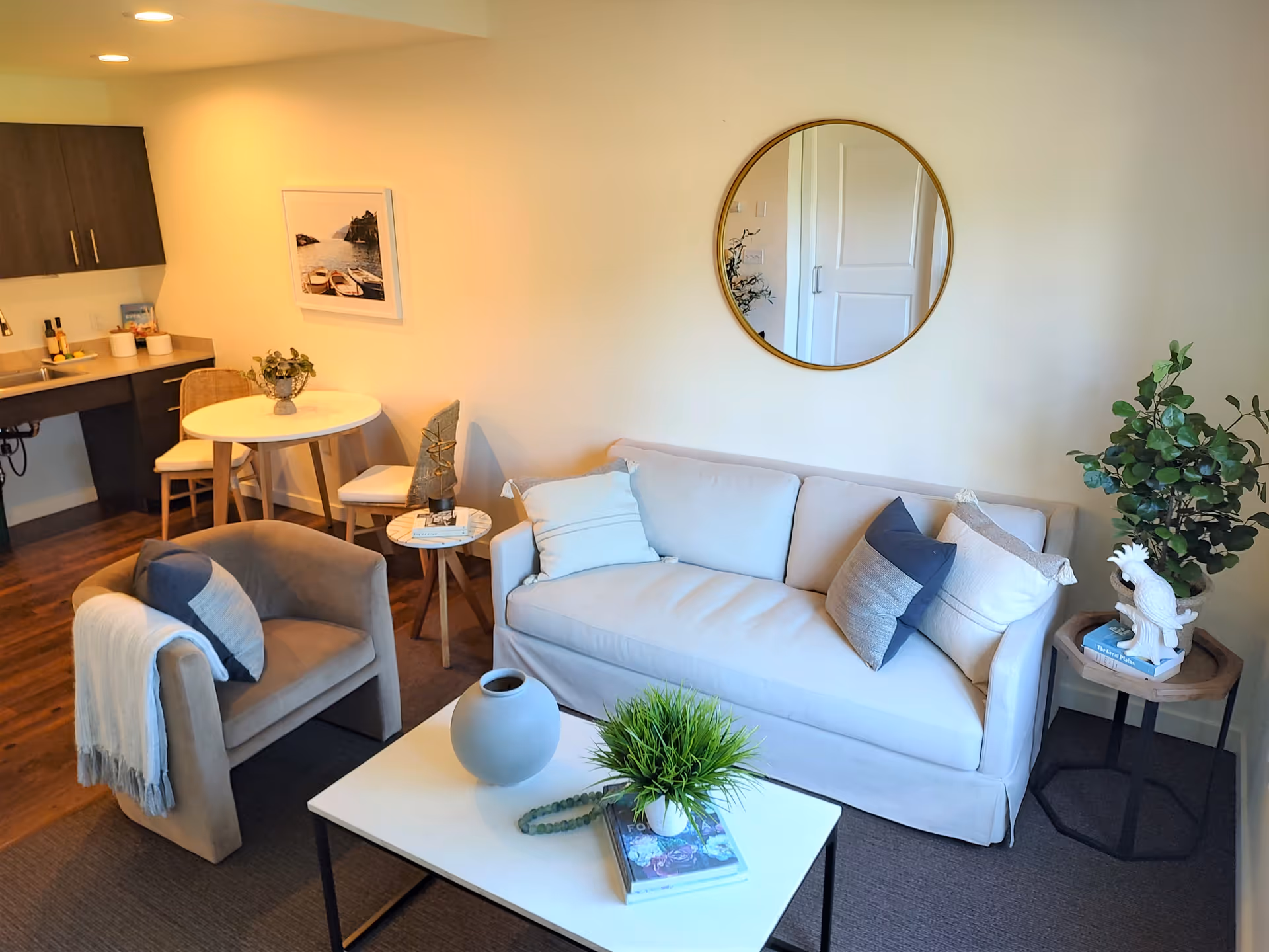 A cozy living room area with a light gray sofa adorned with multiple cushions, a round mirror on the wall above it, a small side table with a decorative white bird statue and a plant, a beige armchair with a throw blanket and pillow, a white coffee table with a gray vase, a green plant, and a book. In the background, there is a small round dining table with two chairs and a kitchenette with dark cabinets and a countertop.