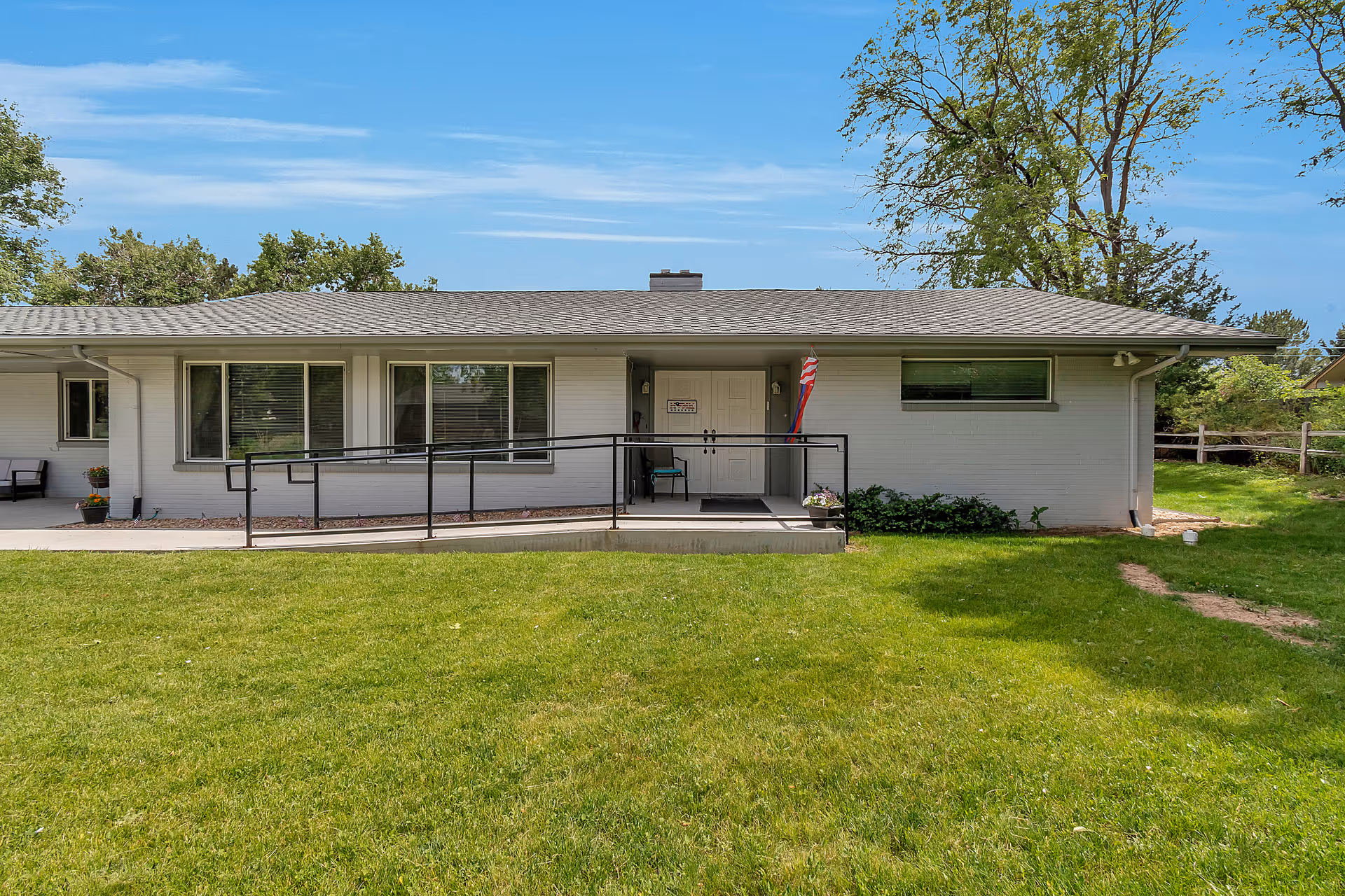 Single-story building with white exterior walls and a gray shingled roof. There is a wheelchair accessible ramp leading to a double door entrance. The building is surrounded by green grass and trees under a blue sky with some clouds.