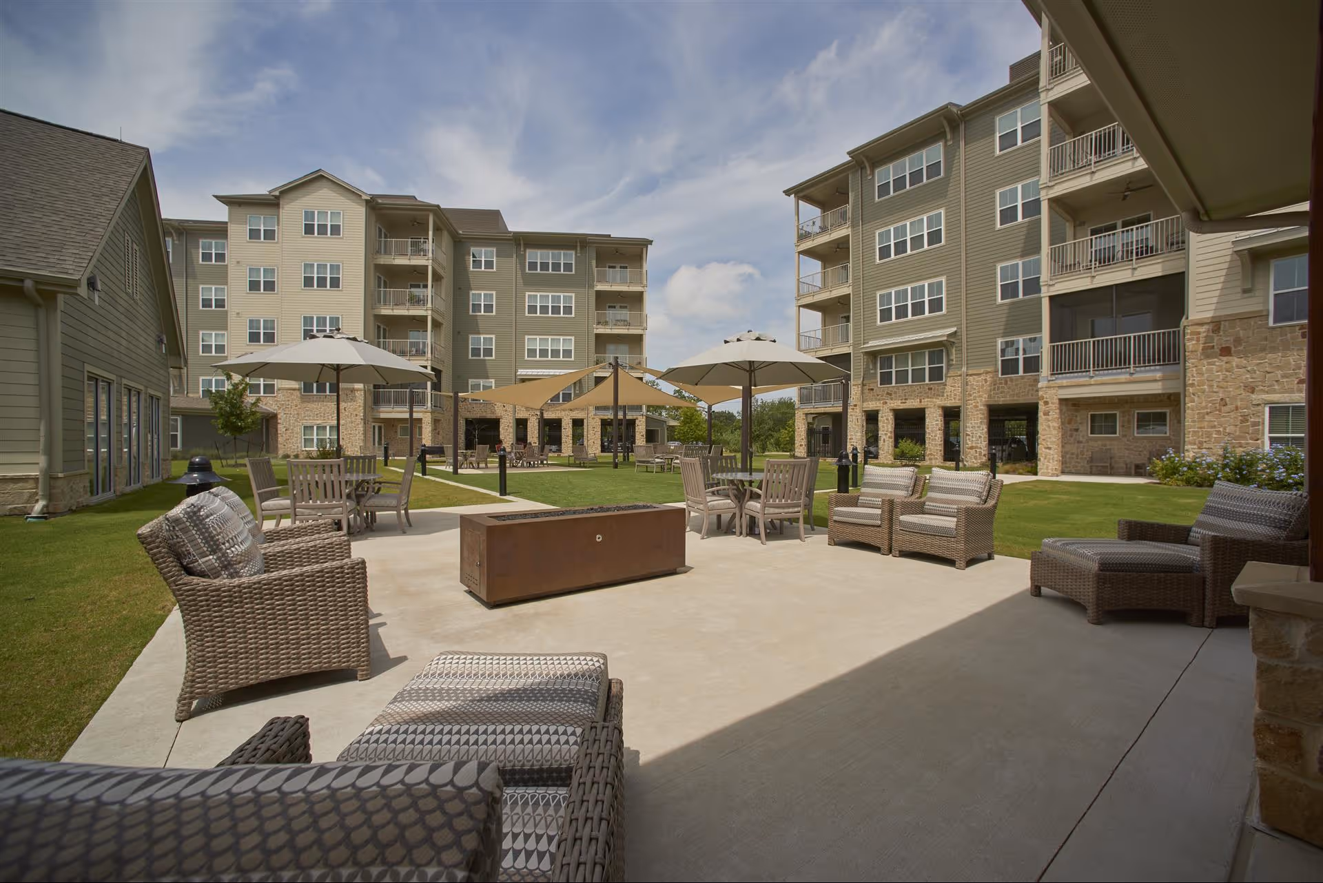 Outdoor patio area at The Langford Methodist Retirement Community featuring wicker lounge chairs, tables with umbrellas, and a fire pit, surrounded by multi-story residential buildings under a partly cloudy sky.